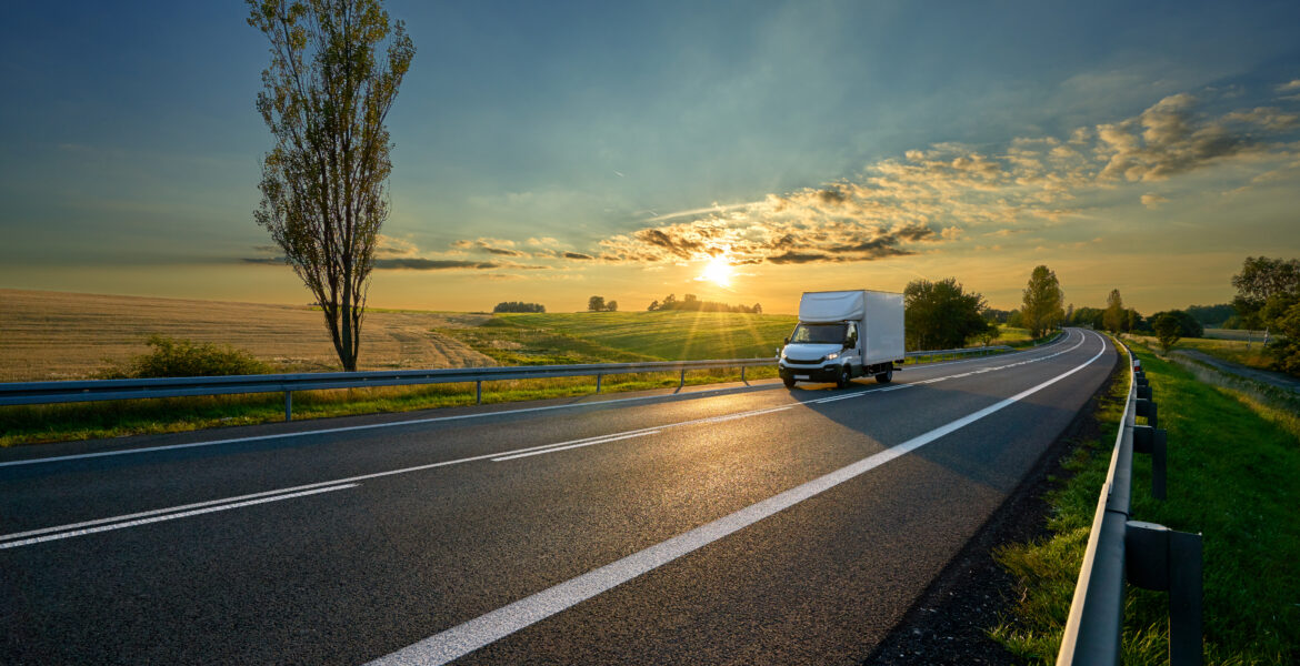 White delivery van driving on asphalt road around farm fields in rural landscape at sunset